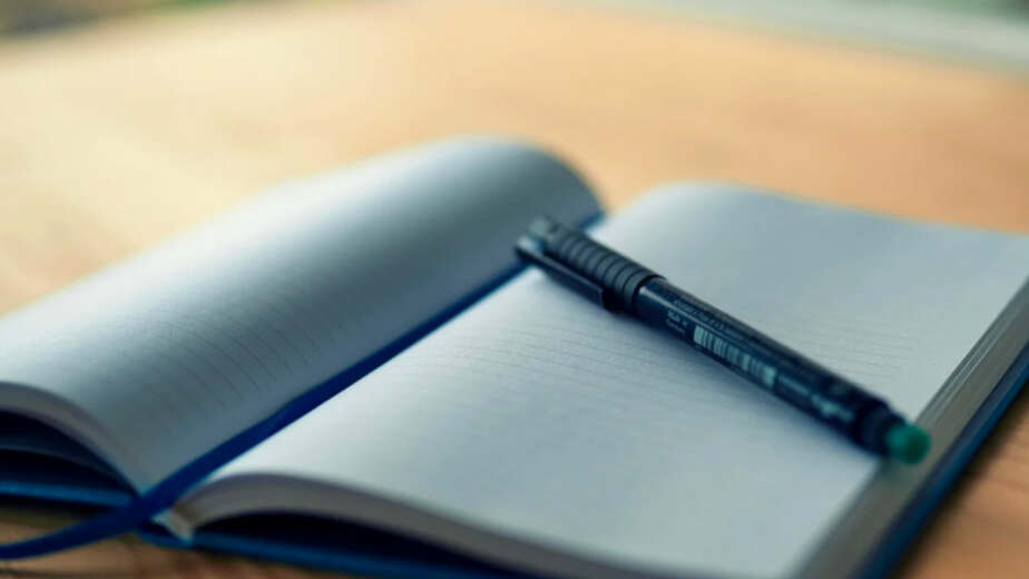 Table en bois, avec un calepin ouvert sur des pages blanches accompagné d'un crayon.