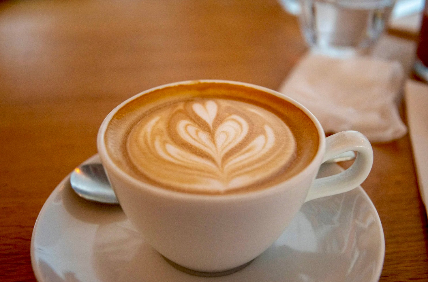 Sur une table, on voit une tasse. À café dans lequel se trouve un latté, sur lequel il y a un motif de cœur, dans la petite assiette on voit une cuillère en argent.